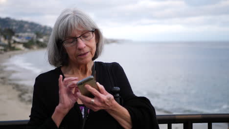 A-middle-aged-woman-with-white-hair-and-eye-glasses-texting-on-her-phone-on-vacation-in-Laguna-Beach,-California-SLOW-MOTION