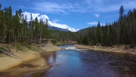 Low-Aerial-view-flying-slow-up-wide-winding-river-over-large-cascade-on-bright-sunny-afternoon-spring-day-in-Colorado-forest-wilderness-with-majestic-snow-capped-mountains,-blue-sky-and-clouds