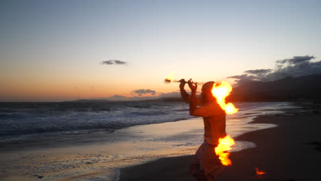 Ein-Mann-In-Silhouette-Tanzt-Und-Dreht-Einen-Brennenden-Feuerstab-Am-Strand-Bei-Sonnenuntergang-Mit-Flammen-Und-Meereswellen-In-Zeitlupe