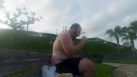 Obese-man-eating-a-greasy-burger-on-a-park-bench-leans-back-then-rubs-his-fat,-hairy-stomach