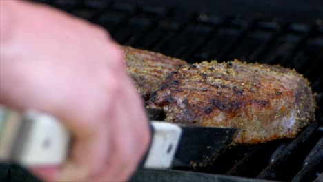 A-pair-of-meat-tongs-turn-a-nearly-cooked-juicy-rib-eye-steak-on-a-grill-and-slides-it-a-bit-in-slow-motion