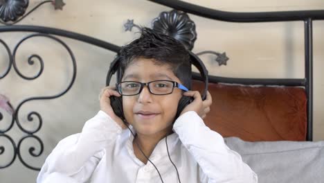 Cute-Little-indian-asian-caucasian-boy-wearing-spectacles-glasses-listening-enjoying-music-on-headphone-hands-on-ear-sitting-in-living-room-looking-at-camera-front-view