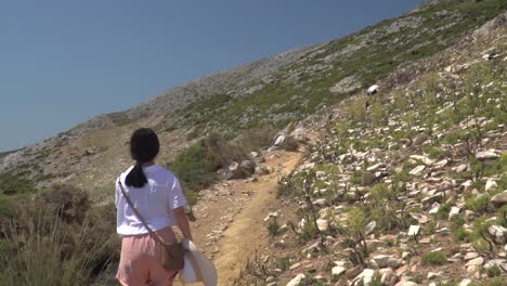 Following-shot-of-girl-in-summery-hiking-outfit-walking-up-path-in-barren-mountain-landscape