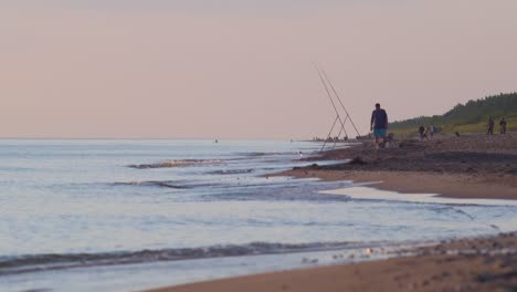 A-fisherman-fishes-for-flatfish-with-a-fishing-rod-on-the-coast-of-Baltic-sea-in-sunny-summer-evening-before-sunset,-wide-shot-from-a-distance