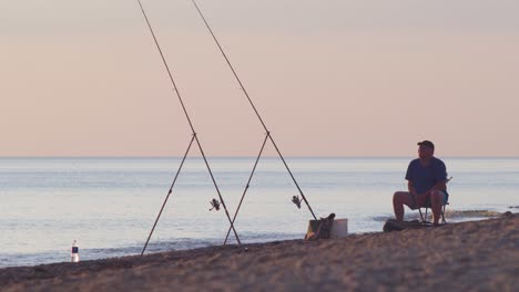 A-fisherman-fishes-for-flatfish-with-a-fishing-rod-on-the-coast-of-Baltic-sea-in-sunny-summer-evening-before-sunset,-medium-shot-from-a-distance