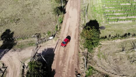 Shot-aerial-drone-footage-tracking-a-red-car-leaving-a-house-in-the-country,-on-an-empty-road-surrounded-by-a-beautiful-forest-trees-a-sunny-day