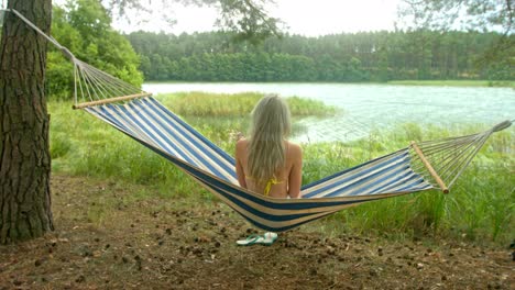 Blond-girl-sits-on-a-hammock-on-a-warm-sunny-summer-day-and-looks-at-the-calm-lake