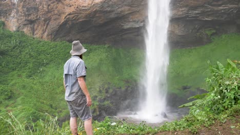 A-bearded-ginger-man-wearing-a-bushman-hat-stands-at-the-base-of-a-powerful-water-fall-in-tropical-Africa