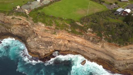 Epic-drone-shot-of-ocean-waves-smashing-against-cliff-and-rocks,-near-a-park,-Sydney,-Australia
