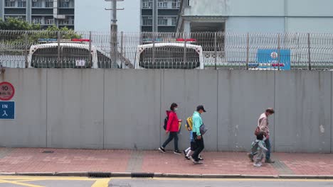 Fahrzeuge-Und-Fußgänger-Fahren-Und-Gehen-An-Einer-Polizeistation-Vorbei,-Die-Durch-Eine-Große,-Hohe-Graue-Mauer-Und-Einen-Stachelzaun-In-Hongkong-Geschützt-Ist