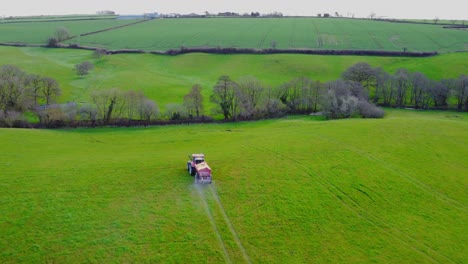 Smooth-aerial-drone-view-following-a-red-tractor-fertilising-a-greet-field-with-trees
