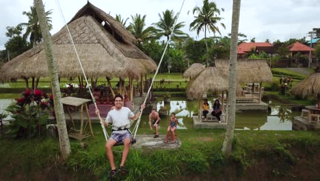 Toma-De-Un-Dron-De-Un-Turista-Disfrutando-De-Un-Columpio-Entre-Dos-Cocoteros-Que-Se-Balancea-Sobre-Algunas-Terrazas-De-Arroz-En-Bali,-Indonesia