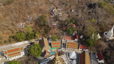 Wat-Phra-Phutthabat,-Saraburi,-Thailand,-reverse-aerial-footage-of-Buddhist-structures-within-the-forest-with-brown-trees-in-summer,-also-revealing-the-golden-temple-and-complex,-road,-green-trees