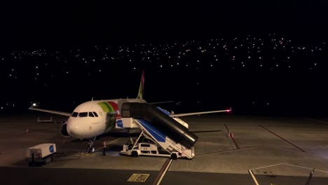 TAP-Night-time-Airport-operations,-After-landing-the-airplane-in-the-dark-a-passenger-boarding-bridge-mobile-staircase-moves-towards-aeroplane-to-support-secure-safety-passenger-offloading