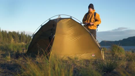 Young-explorer-standing-beside-his-tent-in-heavy-winds-surrounded-by-amazing-untouched-Scandinavian-nature-during-sunset-in-Lappland,-Sweden