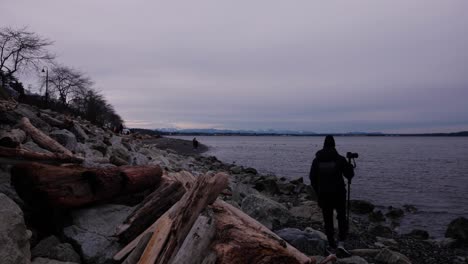 Man-walking-with-tripod-and-camera-over-sandy-rock-beach-covered-in-drift-wood-and-boulders-with-people-walking-in-the-distance-clouds-trees-white-rock-park