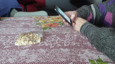 Weiße-Frau-Isst-Erdnüsse-Auf-Einem-Tisch-Und-Benutzt-Handy