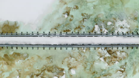Person,-Die-Im-Winter-Auf-Einer-Schmalen-Brücke-über-Einen-Zugefrorenen-Fluss-In-Norwegen-Läuft---Luftaufnahme