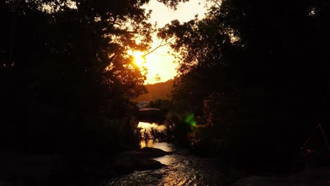 Sunset-over-serene-stream,-golden-light-shining-on-rocks-under-shade-of-trees