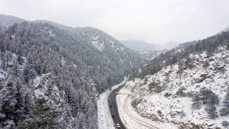 Aerial-reveal-backwards-of-Boulder-Canyon-Drive-in-Colorado-during-the-winter-as-cars-drive-down-icy-road-surrounded-by-rocky-mountains-and-snow-covered-pine-trees
