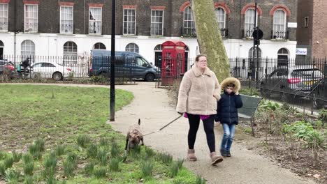 Mother-walking-with-her-child-and-her-dog-in-a-park-in-London-with-a-telephone-red-booth-behind-them