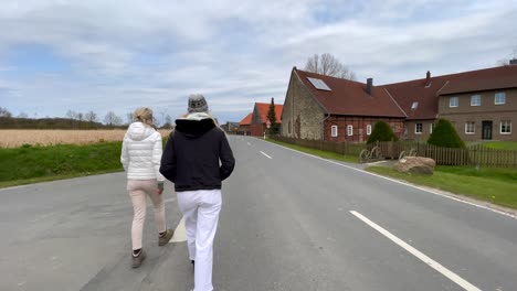 Slow-motion-tracking-shot-of-two-adult-girls-walking-along-road-next-to-rural-wheat-field-and-farm-during-sunny-day-with-clouds