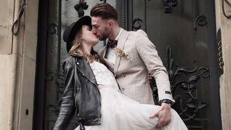 Retro-styled-wedding-couple-with-kissing-passionate-in-front-of-baroque-door---bride-in-black-leather-jacket-and-hat-slowmotion