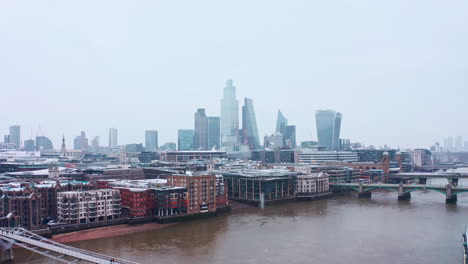 Rising-London-aerial-drone-shot-of-snow-falling-on-city-of-London-skyscrapers-from-river-thames