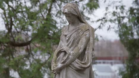 Alte-Traurig-Aussehende-Skulptur-Aus-Stein-Der-Mutter-Maria-Auf-Einem-Alten-Friedhof-Mit-Bäumen,-Die-Im-Wind-Wehen