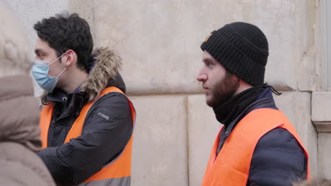 Un-Par-De-Hombres-De-Seguridad-Con-Máscara-Y-Chaqueta-Reflectante-Naranja-En-Guardia-Durante-Una-Protesta-Laboral-En-Milán