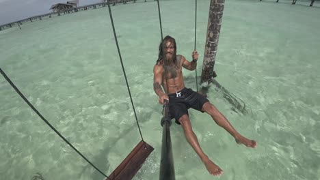 A-young,-fit-and-strong-man-is-sitting-by-himself-on-a-swing,-smiling-with-his-feet-and-legs-in-the-warm-and-clear-water