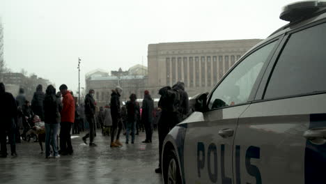 Plano-General-De-Un-Coche-De-Policía-De-Helsinki-Estacionado-Con-Una-Multitud-De-Personas-En-El-Fondo-De-Las-Manifestaciones-De-Covid-19