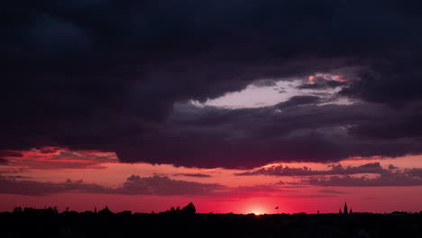 Time-lapse-of-beautiful-scenic-red-sunset-over-the-city-with-sun-rays-shining-through-the-clouds,-high-contrast-low-fast-moving-clouds-at-summer-evening,-urban-environment,-wide-shot