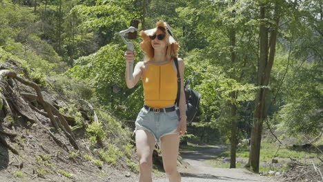 Young-female-vlogger-walking-on-a-hiking-trail-and-recording-vertical-video-with-smartphone-and-mobile-gimbal-on-sunny-day