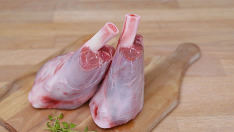 Two-Lamb-Shanks-With-Green-Herbs-On-Wooden-Chopping-Board