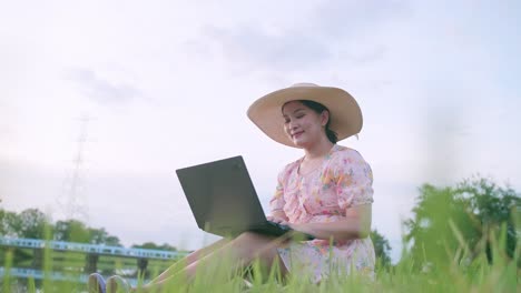 Beautiful-Asian-woman-in-hat-sitting-on-lawn-working-on-computer-feeling-successful