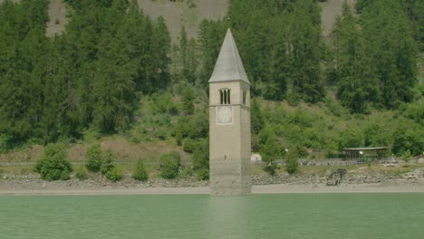 Full-shot,-scenic-view-of-Kirchturm-von-Altgraun-in-Italy,-Reschensee-shore-and-forest-range-in-the-background