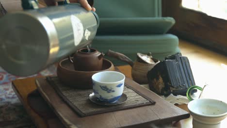 Water-being-poured-from-a-thermos-into-a-clay-teapot-on-a-tea-table-next-to-other-tea-ware