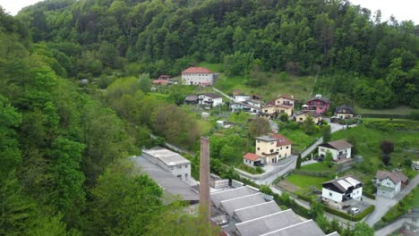 Drone-going-up-in-front-of-small-factory-chimney-with-settlement-between-green-nature-in-back
