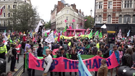 Hundreds-of-colourful-Extinction-Rebellion-climate-change-protesters-occupy-a-Covent-Garden-road-junction-and-set-up-a-giant-pink-table-blocking-the-road-behind-a-pink-banner-saying-“Stop-the-harm”