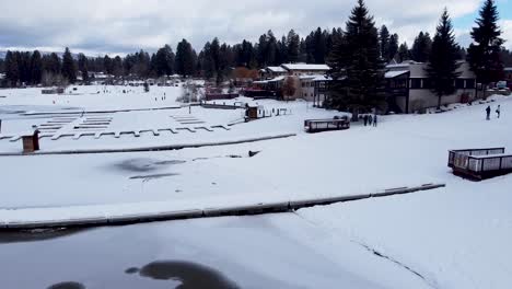 Snowy-icy-frozen-lake-in-winter-and-empty-snow-covered-boat-wharf-piers,-pine-tress-and-small-town-in-McCall,-Idaho,-USA---Aerial-drone-towards-town