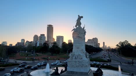 Aerial-slow-pull-out-shot-capturing-monumental-sculpture-Carta-Magna-and-Four-Regions-of-Argentina-at-rotary-road-with-peak-hour-traffics-moving-and-downtown-cityscape-in-the-background