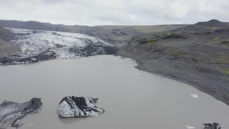 Aerial-view-over-the-Solheimajokull-glacier-lagoon,-in-cloudy-Iceland---reverse,-drone-shot