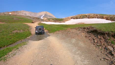 POV-following-a-4WD-vehicle-going-up-Black-Bear-Pass-trail,-past-grass-covered-hills-and-dirty-snow-drifts-in-San-Juan-Mountains-near-Telluride-Colorado