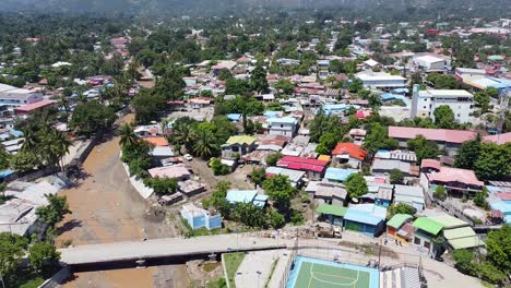 Aerial-drone-of-capital-city-Dili-after-natural-disaster-cyclone-and-flash-floods-in-Timor-Leste,-South-East-Asia,-lowering-over-buildings,-river-and-football-pitch