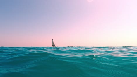 Low-angle-sea-level-view-of-small-sailboat-sailing-in-calm-open-turquoise-sea-water-with-pink-sky-in-background