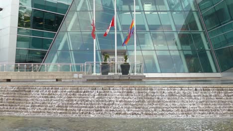 Canada-and-rainbow-flag-waving-near-modern-building,-handheld-view
