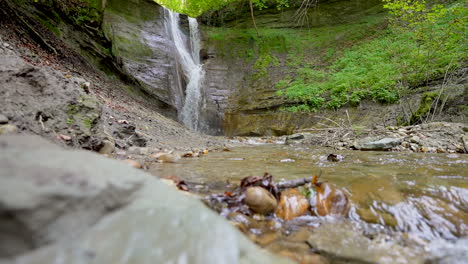 Low-angle-shot-of-slowly-flowing-natural-creek-and-waterfall-in-background-surrounded-by-woodland-during-sunny-day---Slow-motion-pan-shot