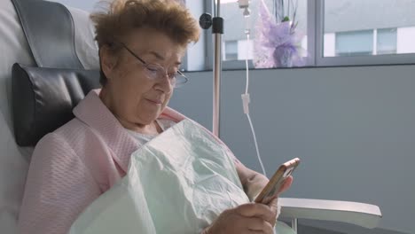 Hospital-room,-Close-up-Elderly-woman-holding-mobile-phone,-intravenous-drip-Medication
