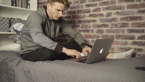 Ein-Junger-Mann-Sitzt-Auf-Seinem-Bett-Und-Tippt-Und-Arbeitet-An-Seinem-MacBook-Pro-Laptop